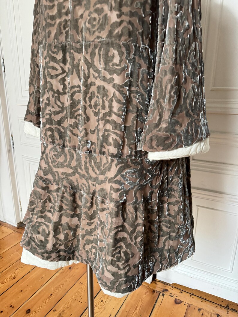 Puede incluir: Vestido vintage en tonos marr&oacute;n y gris con estampado floral. Mangas largas con pu&ntilde;os blancos y falda escalonada con ribete blanco. La tela parece terciopelo. El vestido se exhibe en un soporte.