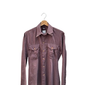 May include: A long-sleeved, button-down shirt with vertical stripes in shades of burgundy and white. The shirt has two chest pockets and is hanging on a wooden hanger. The shirt has a classic, casual style.