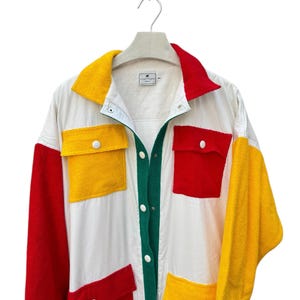 Puede incluir: Una camisa blanca de manga larga con botones con paneles rojos, amarillos y verdes. La camisa tiene bolsillos grandes en la parte delantera y un cuello de contraste.