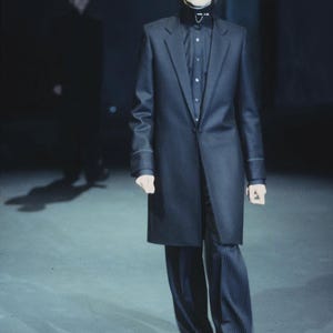 May include: A long black coat worn over a black button-down shirt and dark pinstriped trousers. The ensemble features a high-collared black shirt with a small silver detail at the neck, paired with a structured coat and loose-fitting pinstriped pants.