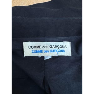 Pode incluir: Pe&ccedil;a de vestu&aacute;rio preta com uma etiqueta branca com o texto "COMME des GAR&Ccedil;ONS" em negrito, sem serifa. A etiqueta est&aacute; centrada e tem um detalhe de estrela azul. O tecido da pe&ccedil;a parece ser um material escuro, possivelmente tecido.