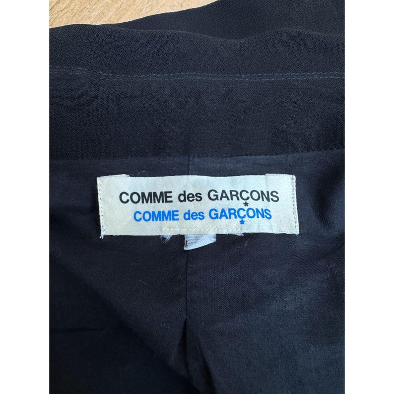 Pode incluir: Pe&ccedil;a de vestu&aacute;rio preta com uma etiqueta branca com o texto "COMME des GAR&Ccedil;ONS" em negrito, sem serifa. A etiqueta est&aacute; centrada e tem um detalhe de estrela azul. O tecido da pe&ccedil;a parece ser um material escuro, possivelmente tecido.