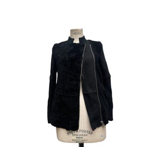 May include: A black jacket with a textured, fuzzy front panel and smooth black sleeves. The jacket features a full-length zipper and is displayed on a vintage dress form. The jacket has a high collar.