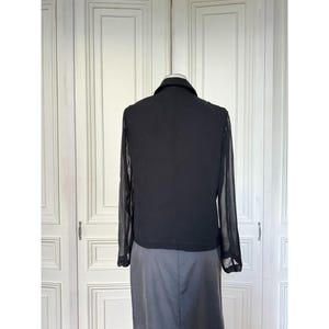 Pode incluir: Blusa preta de manga comprida com gola e mangas transparentes. A blusa &eacute; combinada com uma saia cinza. A roupa &eacute; exibida em um manequim em um fundo branco.