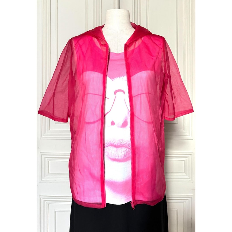 May include: A bright pink, short-sleeved, hooded jacket over a pink graphic t-shirt. The jacket is sheer and has a zipper closure. The t-shirt features a graphic of a face. The bottom of the outfit is a black skirt.