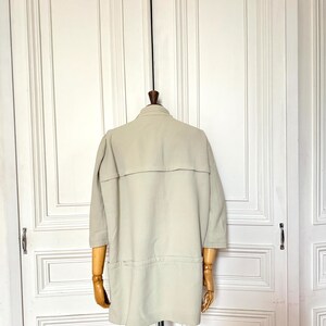 Puede incluir: Una chaqueta beige claro con mangas tres cuartos cuelga de un maniqu&iacute; de madera. La chaqueta tiene cuello, dos bolsillos y una costura horizontal en la espalda. El fondo es una puerta blanca.