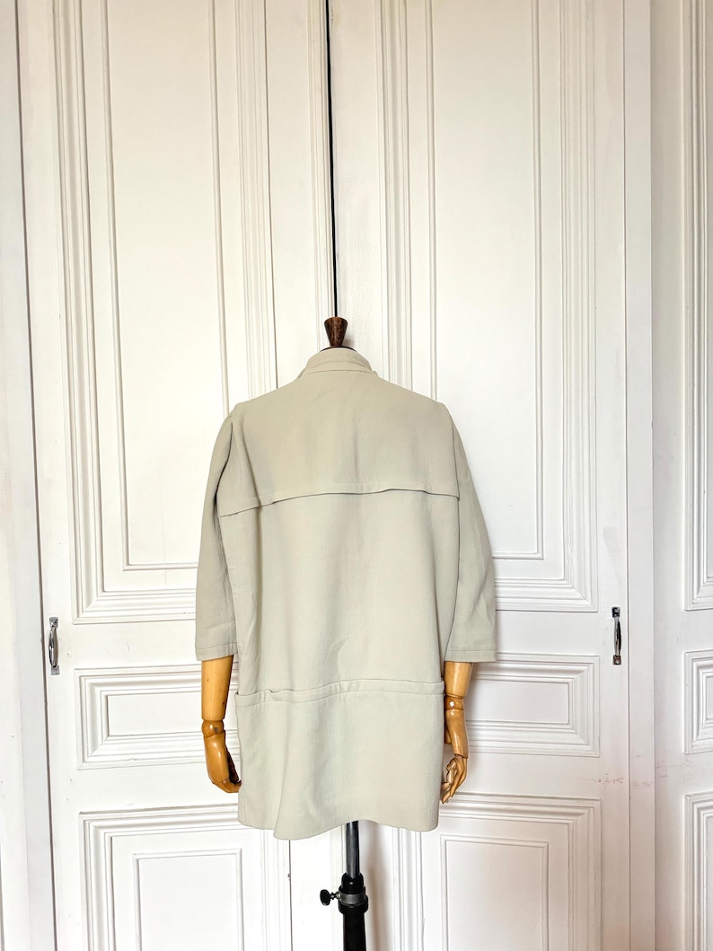 Puede incluir: Una chaqueta beige claro con mangas tres cuartos cuelga de un maniqu&iacute; de madera. La chaqueta tiene cuello, dos bolsillos y una costura horizontal en la espalda. El fondo es una puerta blanca.