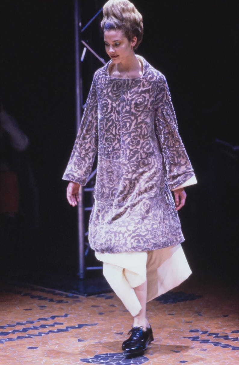 Puede incluir: Un vestido largo de manga larga, hasta la rodilla, con un estampado floral en tonos grises y morados. El vestido se combina con pantalones color crema y zapatos negros. La modelo est&aacute; sobre un suelo de madera.