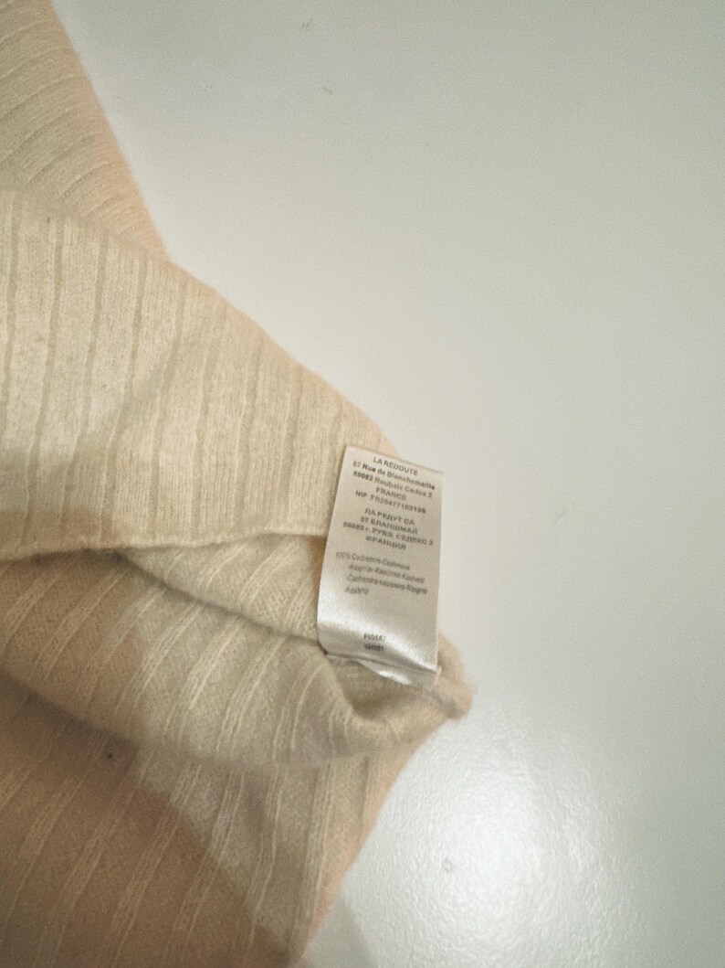 May include: A beige and cream colored knitted sweater with a tag that reads "LA REDOUTE" and includes information about the fabric and care instructions.