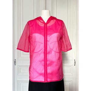 May include: A bright pink, sheer hooded jacket with short sleeves and a full-length zipper. The jacket is displayed on a mannequin, revealing a lighter pink underlayer. The garment is lightweight and appears to be made of a delicate fabric.
