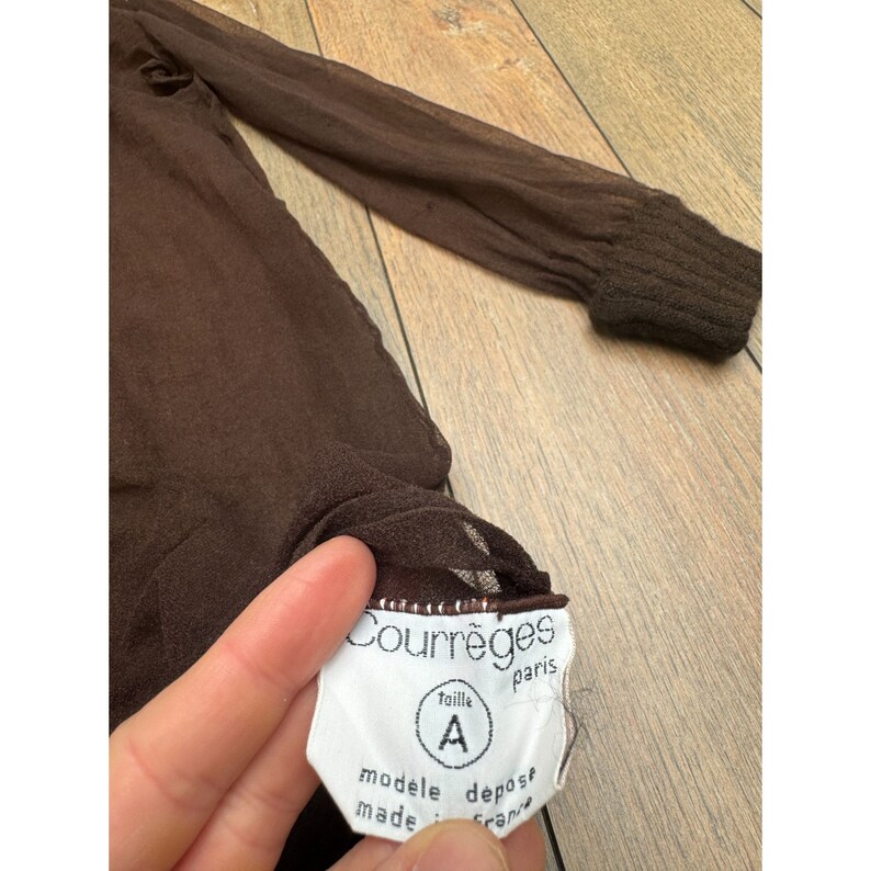 Op de afbeelding: Bruin doorschijnend lang mouw shirt met een wit label waarop "Courr&egrave;ges Paris" en "taille A modele depose made in France" staat.
