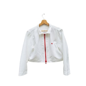 Puede incluir: Una chaqueta blanca corta con una cremallera roja y un peque&ntilde;o logotipo rojo en el lado derecho. La chaqueta est&aacute; colgada en una percha de madera.