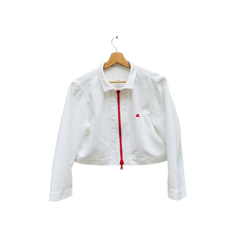 Puede incluir: Una chaqueta blanca corta con una cremallera roja y un peque&ntilde;o logotipo rojo en el lado derecho. La chaqueta est&aacute; colgada en una percha de madera.