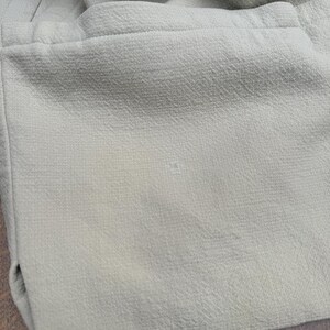 Puede incluir: Primer plano de una tela verde claro con superficie texturizada. La tela parece ser parte de una prenda, con costuras visibles y un detalle de bolsillo. El color es un verde p&aacute;lido y apagado.