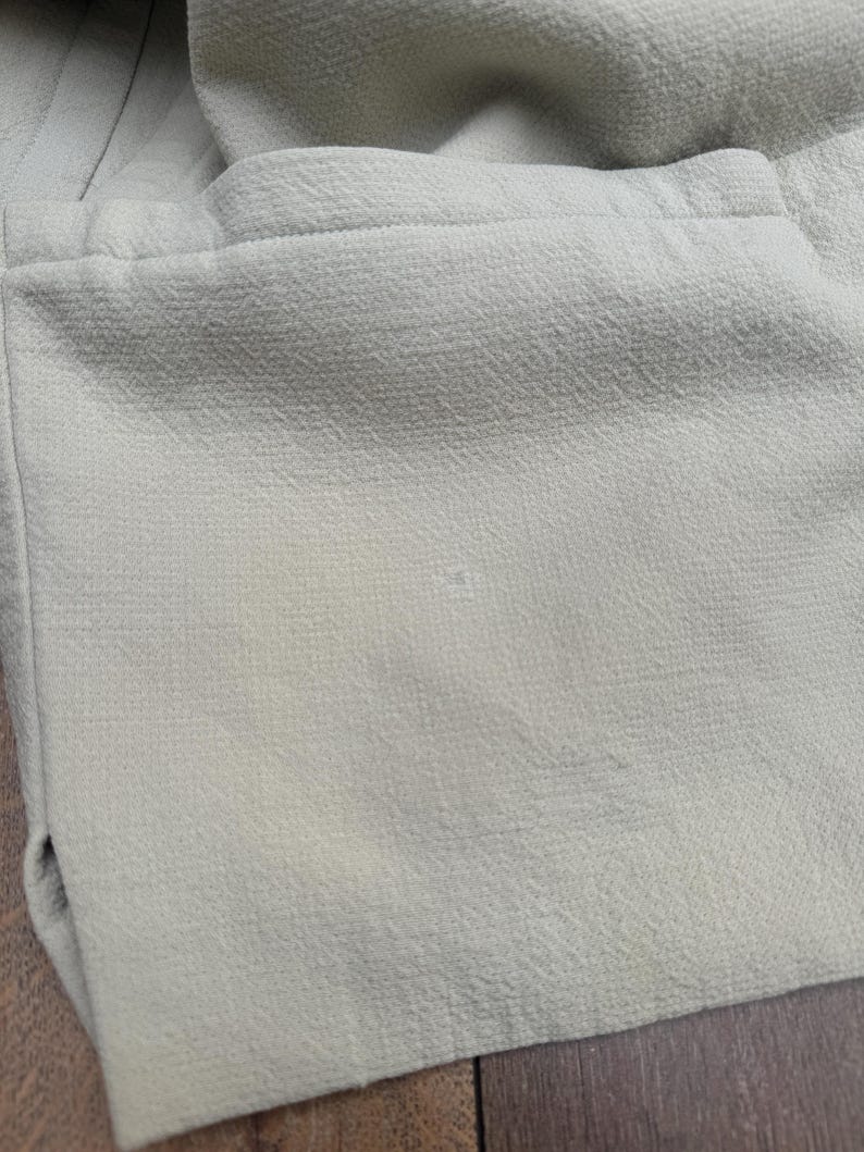 Puede incluir: Primer plano de una tela verde claro con superficie texturizada. La tela parece ser parte de una prenda, con costuras visibles y un detalle de bolsillo. El color es un verde p&aacute;lido y apagado.