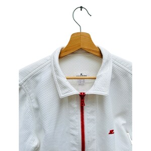 Puede incluir: Chaqueta acolchada blanca con cremallera roja y un peque&ntilde;o logotipo bordado en el pecho. La chaqueta est&aacute; colgada en una percha de madera.