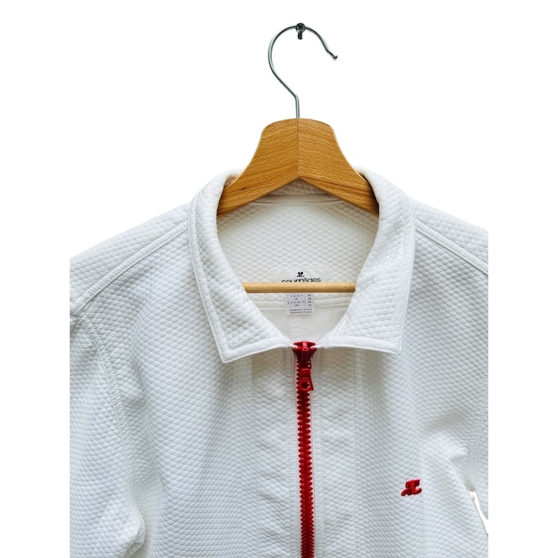 Puede incluir: Chaqueta acolchada blanca con cremallera roja y un peque&ntilde;o logotipo bordado en el pecho. La chaqueta est&aacute; colgada en una percha de madera.