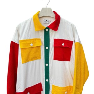 Puede incluir: Una camisa blanca de manga larga con botones, una tapeta de botones verde y grandes paneles de patchwork rojo, amarillo y verde. La camisa tiene grandes bolsillos de parche en la parte delantera.