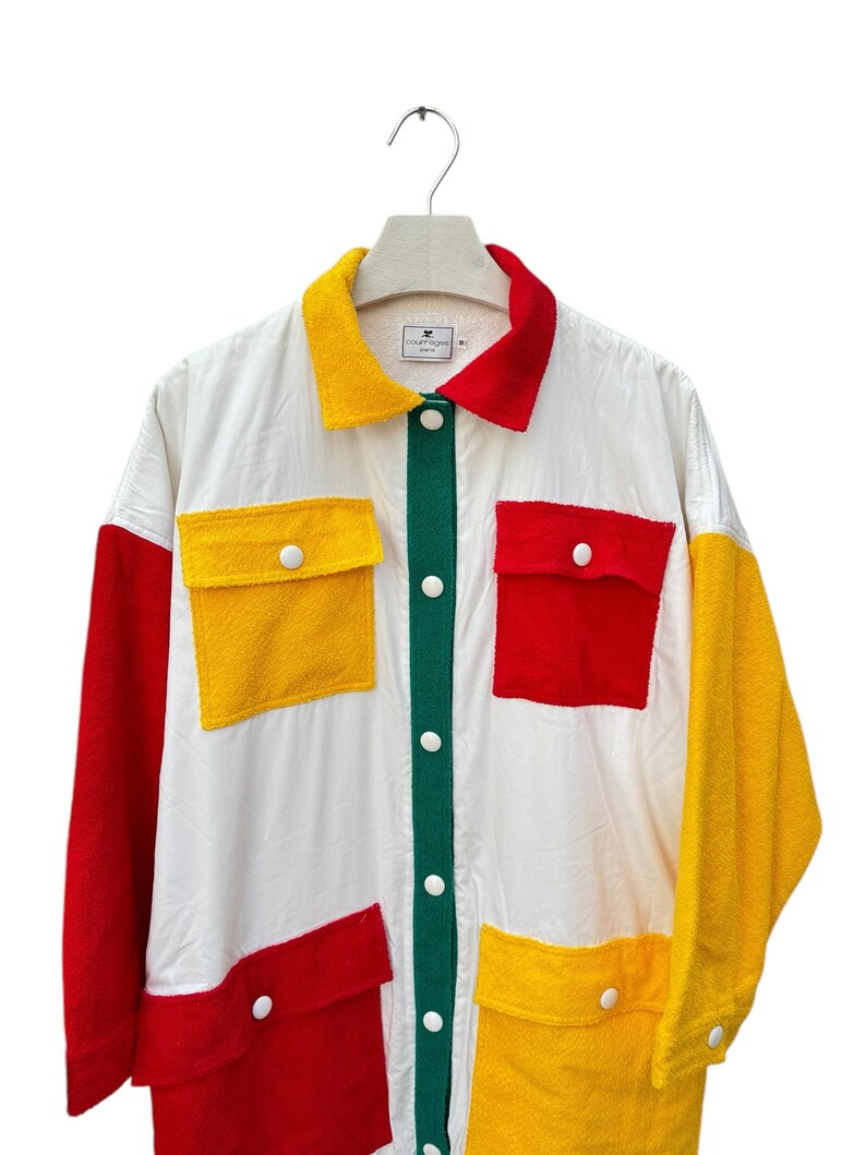 Puede incluir: Una camisa blanca de manga larga con botones, una tapeta de botones verde y grandes paneles de patchwork rojo, amarillo y verde. La camisa tiene grandes bolsillos de parche en la parte delantera.