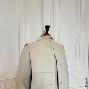 Puede incluir: Un abrigo beige claro con cuello alto y cierre de botones. El abrigo presenta un dise&ntilde;o &uacute;nico tipo capa sobre los hombros y botones azul claro. El abrigo se exhibe en un maniqu&iacute; de madera.