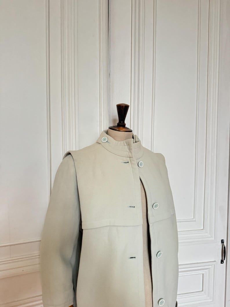 Puede incluir: Un abrigo beige claro con cuello alto y cierre de botones. El abrigo presenta un dise&ntilde;o &uacute;nico tipo capa sobre los hombros y botones azul claro. El abrigo se exhibe en un maniqu&iacute; de madera.