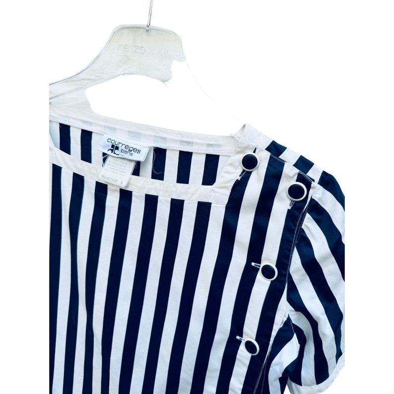 May include: A white and black striped short-sleeved shirt with black buttons. The shirt has a round neckline and the label "Courr&egrave;ges" is visible on the front.