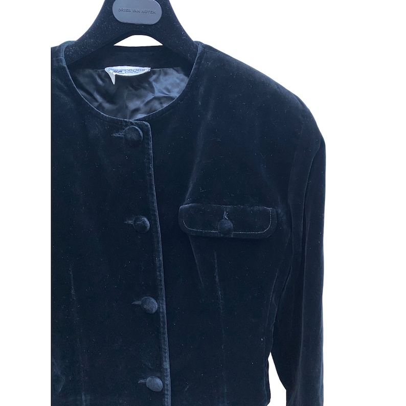 May include: A black velvet jacket with a round neckline, long sleeves, and a single button closure. The jacket has a single pocket on the left side.