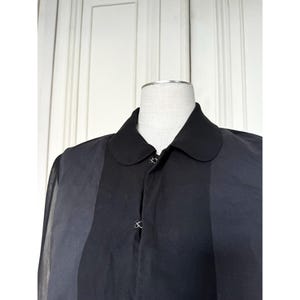 Pode incluir: Uma camisa preta com gola de design &uacute;nico. A camisa apresenta uma faixa vertical cinza escuro no lado esquerdo e uma gola preta. A camisa &eacute; abotoada com pequenos bot&otilde;es decorativos.