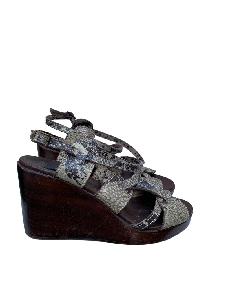 May include: A pair of wedge sandals with a snakeskin pattern in shades of gray, brown, and black. The sandals have a wooden wedge heel and multiple straps that cross over the foot and ankle. The heel is approximately 4 inches high.