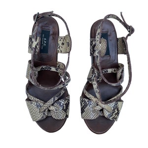 May include: A pair of high-heeled sandals with a snakeskin pattern in shades of brown, tan, and black. The sandals have multiple straps and a buckle closure. The brand name "A.P.C." is visible on the insole.