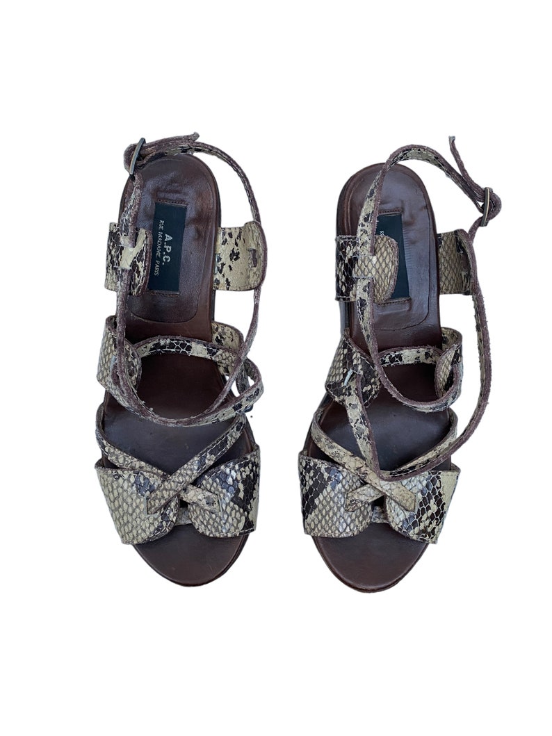 May include: A pair of high-heeled sandals with a snakeskin pattern in shades of brown, tan, and black. The sandals have multiple straps and a buckle closure. The brand name "A.P.C." is visible on the insole.