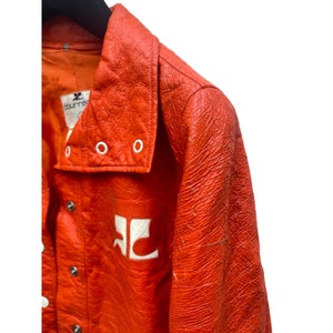 May include: A bright orange leather jacket with a white logo on the front. The jacket has a large collar with white eyelets and a button closure.