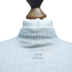 May include: A light gray turtleneck sweater with a small embroidered logo on the front. The logo is a stylized "JC" in white.