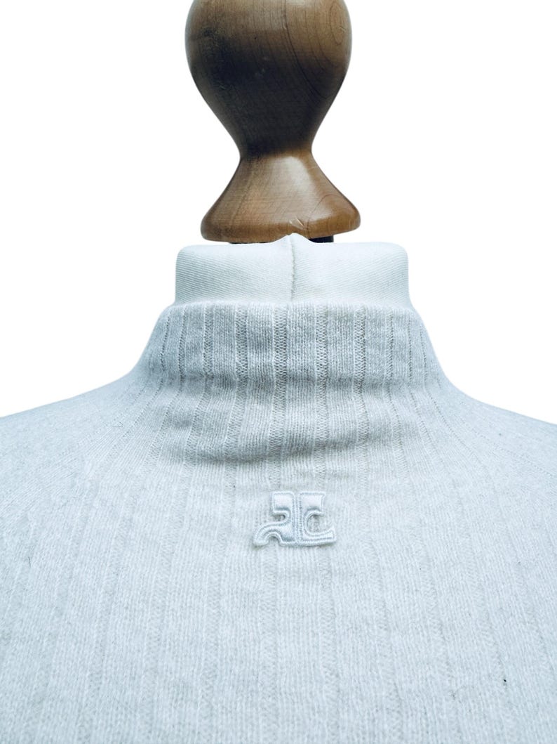May include: A light gray turtleneck sweater with a small embroidered logo on the front. The logo is a stylized "JC" in white.