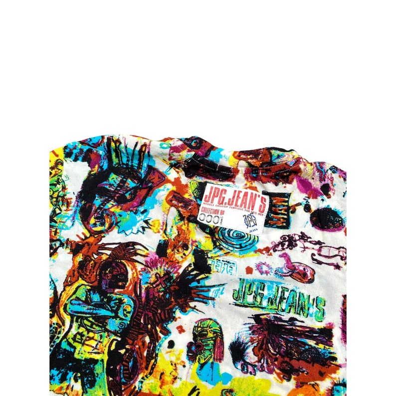 Może przedstawiać: Biała koszula z żywym, kolorowym nadrukiem przedstawiającym abstrakcyjne figury, instrumenty muzyczne i napis "JPG JEAN'S". Projekt zawiera odcienie niebieskiego, ż&oacute;łtego, czerwonego i czarnego.