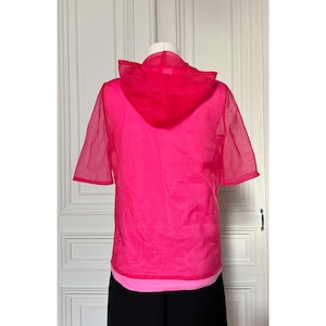 May include: A bright pink, short-sleeved hooded top. The top is made of a sheer material, with a solid pink hood and a lighter pink layer visible at the hem. The garment is displayed on a mannequin against a white door.