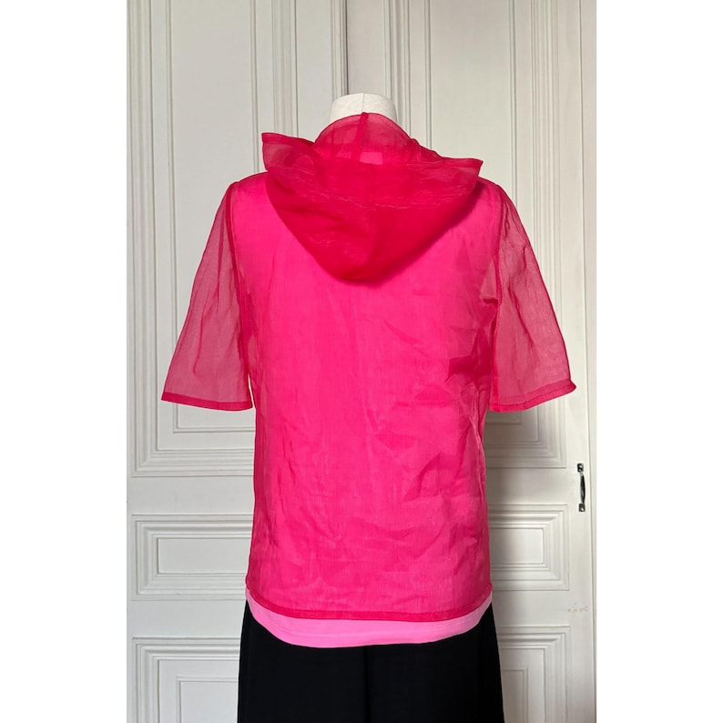 May include: A bright pink, short-sleeved hooded top. The top is made of a sheer material, with a solid pink hood and a lighter pink layer visible at the hem. The garment is displayed on a mannequin against a white door.