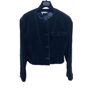 May include: A black velvet cropped jacket with a round neckline, four buttons down the front, and a single pocket on the left side.