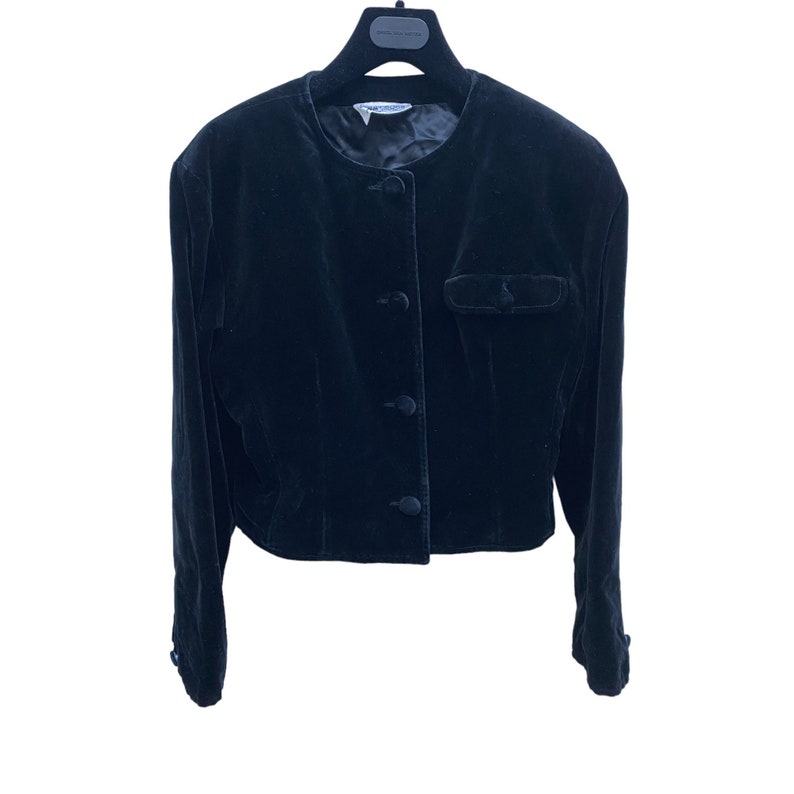 May include: A black velvet cropped jacket with a round neckline, four buttons down the front, and a single pocket on the left side.