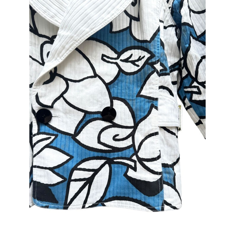 K&ouml;nnte beinhalten: Eine wei&szlig;e Jacke mit blauem Blumendruck, doppelreihigem Verschluss und gro&szlig;em Kragen. Die Jacke hat einen G&uuml;rtel an der Seite.