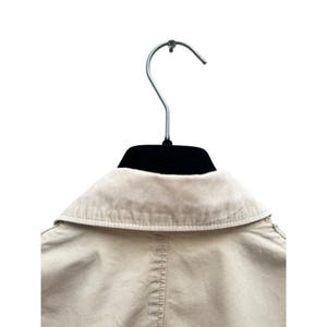 May include: A beige cotton jacket with a collared neckline and a button closure. The jacket is hanging on a black hanger.
