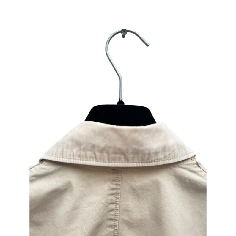 May include: A beige cotton jacket with a collared neckline and a button closure. The jacket is hanging on a black hanger.