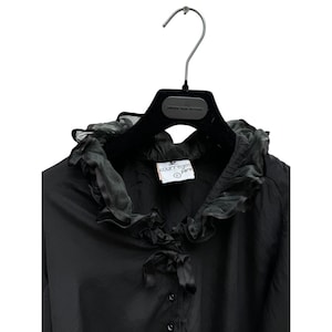 May include: Black long-sleeve blouse with a large ruffled collar and a bow tie. The blouse has a button closure and a label that reads "JC Courr&egrave;ges Paris".