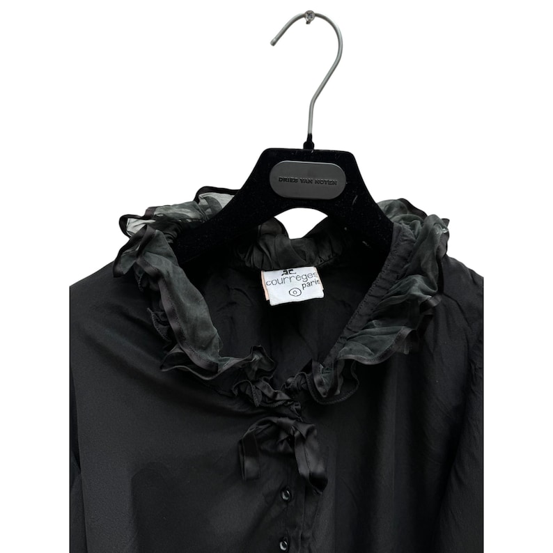 May include: Black long-sleeve blouse with a large ruffled collar and a bow tie. The blouse has a button closure and a label that reads "JC Courr&egrave;ges Paris".
