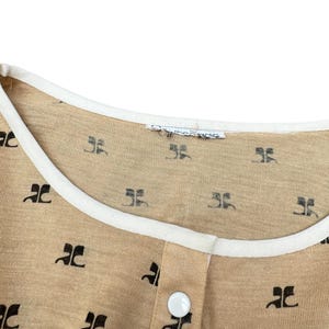 May include: A beige shirt with a white trim around the neckline. The shirt has a small button closure and is covered in a repeating pattern of black "JC" logos.