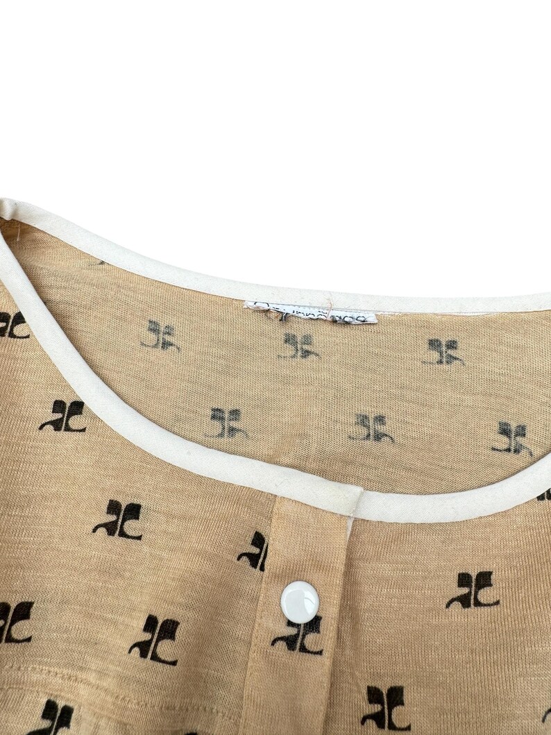 May include: A beige shirt with a white trim around the neckline. The shirt has a small button closure and is covered in a repeating pattern of black "JC" logos.