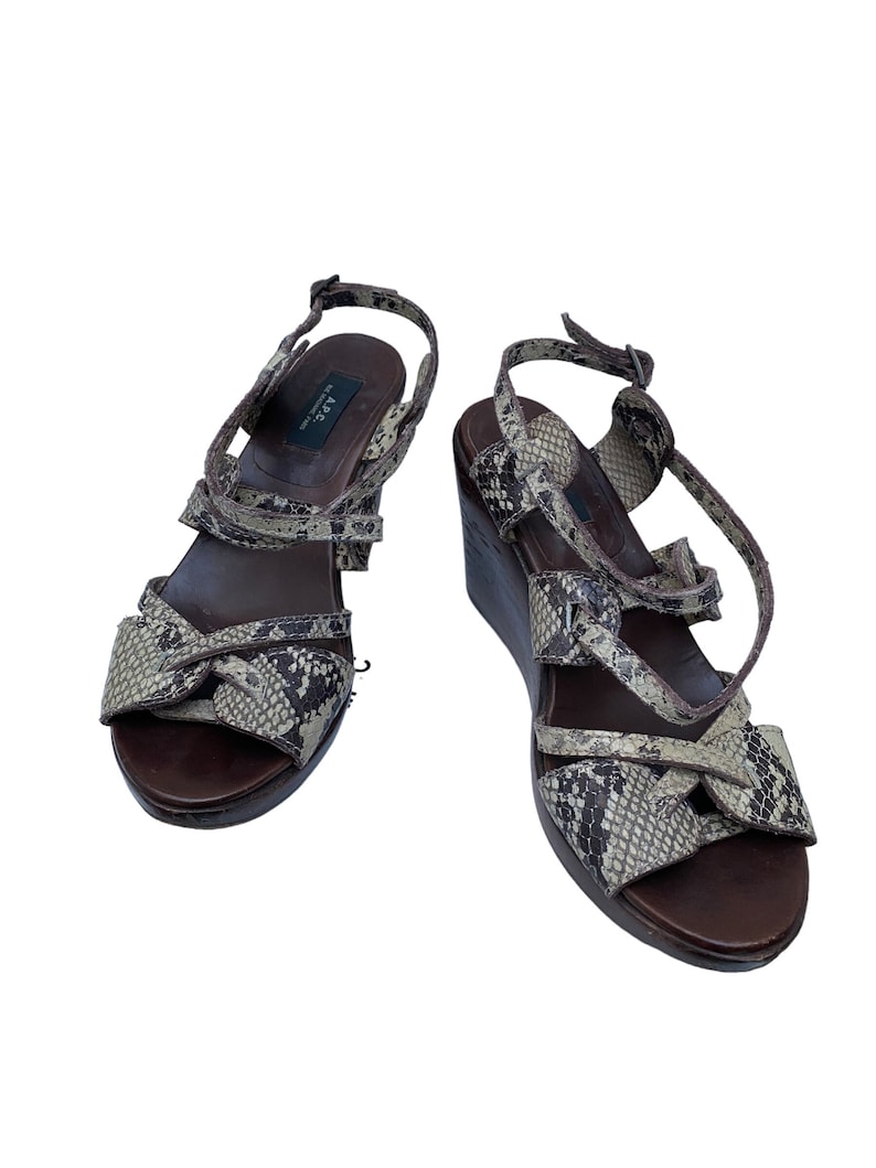 May include: A pair of wedge sandals with a snakeskin pattern in shades of brown, tan, and black. The sandals have a dark brown wooden sole and a buckle closure. The brand name "A.P.C." is visible on the inner sole.