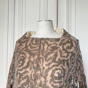 Puede incluir: Una capa de estilo vintage con un estampado de rosas en tonos marr&oacute;n y beige, exhibida en un maniqu&iacute;. La capa presenta un escote cuadrado y una tela suave y fluida. El fondo es una puerta blanca. El dise&ntilde;o de la capa sugiere lujo.