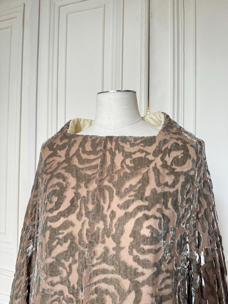 Puede incluir: Una capa de estilo vintage con un estampado de rosas en tonos marr&oacute;n y beige, exhibida en un maniqu&iacute;. La capa presenta un escote cuadrado y una tela suave y fluida. El fondo es una puerta blanca. El dise&ntilde;o de la capa sugiere lujo.