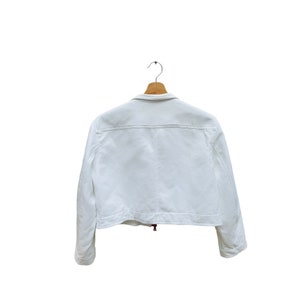 Puede incluir: Chaqueta blanca recortada con cierre de cremallera y mangas largas. La chaqueta est&aacute; colgada en una percha de madera.